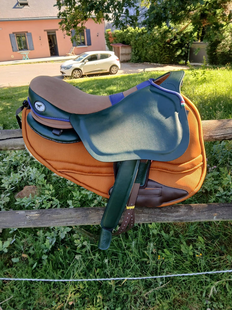 Sella treeless Ghost personalizzata in verde scuro, marrone e arancione, posizionata su una recinzione di legno all'aperto con una casa e un'auto bianca sullo sfondo.