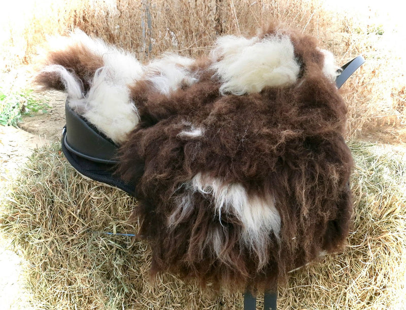 Sella equestre modello Ghost Mandriana Rotondo ricoperta in pelo lungo/montone selvaggio (wild sheepskin) con motivo pezzato bianco e marrone scuro, appoggiata su una balla di fieno all'aperto.